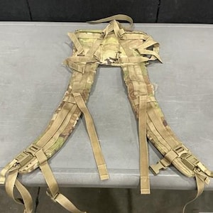 May include: A camouflage patterned backpack harness system featuring two padded shoulder straps, adjustable tan nylon webbing straps, and plastic quick-release buckles. The harness includes a rectangular back panel with integrated attachment points.
