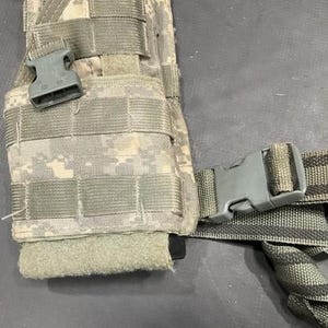 May include: A camouflage tactical vest with a black and gray strap and two black plastic buckles.