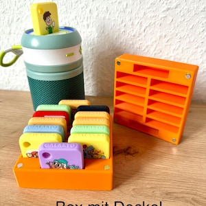 Könnte beinhalten: Orangefarbene Plastikbox mit Deckel, die kleine, farbige quadratische Kacheln mit Comicfiguren und dem Wort "GALAKTO" darauf enthält. Die Box steht auf einer Holzoberfläche. "Box mit Deckel" steht unter der Box.