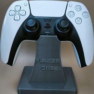 Può includere: Un controller bianco e nero per PlayStation 5 appoggiato su un supporto grigio scuro. Il supporto reca la scritta "PLAYER ONE". Il controller presenta un design simmetrico con joystick neri e pulsanti di azione. Il logo PlayStation è visibile.