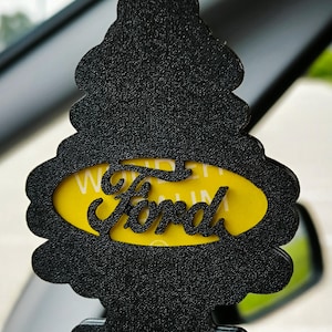 Pode incluir: Ambientador preto em forma de pinheiro com o logotipo da Ford num oval amarelo. O ambientador está pendurado por um fio amarelo, visível no interior de um carro.