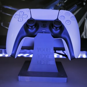 Op de afbeelding: Een wit en zwarte gamecontroller die op een blauwe standaard rust. De standaard heeft de tekst "YOUR TEXT HERE" gegraveerd. De controller heeft een symmetrisch ontwerp met verschillende knopsymbolen.