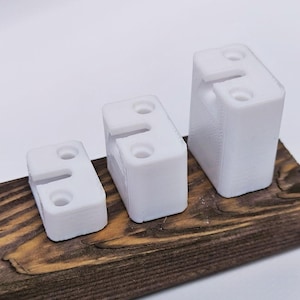 Könnte beinhalten: Drei weiße, rechteckige 3D-gedruckte Objekte unterschiedlicher Größe sind in einer Reihe angeordnet. Jedes Objekt hat einen Schlitz und zwei kreisförmige Löcher. Sie sind auf einer dunkelbraunen Holzoberfläche vor weißem Hintergrund platziert.