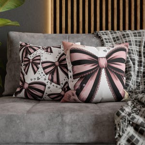 May include: Two decorative throw pillows with pink and black bow designs on a white background. One pillow features multiple bows, hearts, and floral accents. The other pillow has a large bow with a scalloped edge. The pillows are on a gray couch.