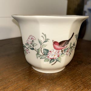 Puede incluir: Jardinera de cerámica color crema con diseño floral y de pájaros. La jardinera presenta un pájaro rosa posado en una rama con flores rosas y hojas verdes. El borde tiene un ribete dorado.