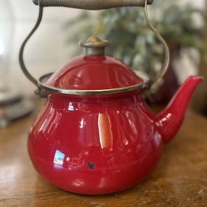 Puede incluir: Una tetera de esmalte rojo vibrante con asa y adornos de latón. La tetera tiene un cuerpo redondeado, un pico curvo y una tapa con un pomo de latón. El asa se arquea sobre la parte superior y la tetera está sobre una superficie de madera.