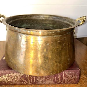 May include: A vintage brass pot with ornate handles. The pot has a rounded body and a wide opening. It sits on a maroon book with gold lettering. The pot's surface shows signs of age and use, adding to its antique charm.