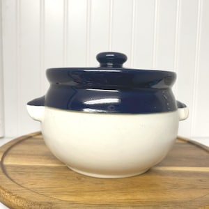 May include: A ceramic crock pot with a lid. The pot is white with a dark blue band around the top and a matching lid. It has two small handles on either side. The crock pot is sitting on a wooden surface.
