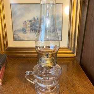 Könnte beinhalten: Eine Öllampe aus klarem Glas mit Griff und hohem, gebogenem Schornstein. Die Lampe hat einen messingfarbenen Brenner und steht auf einer Holzoberfläche. Der Hintergrund umfasst ein gerahmtes Gemälde mit einem goldenen Rahmen.