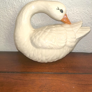 May include: A cream-coloured ceramic goose figurine with an orange beak and black eye. The goose is in a curved pose, with its head bent towards its back. The figurine is placed on a wooden surface.