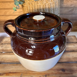 May include: A vintage brown and cream-colored stoneware crock pot with a lid and two handles. The crock pot has a glossy finish and a classic design, perfect for kitchen decor or cooking. The lid features a small, round knob.