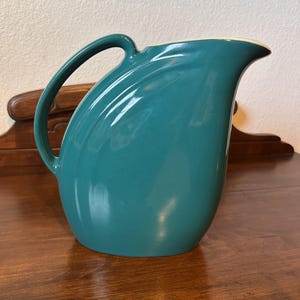 Op de afbeelding: Teal keramische kan met een gebogen handvat en tuit. De kan heeft een gladde, glanzende afwerking en een crèmekleurige rand. Het ontwerp is eenvoudig en elegant, geschikt voor het serveren van dranken.