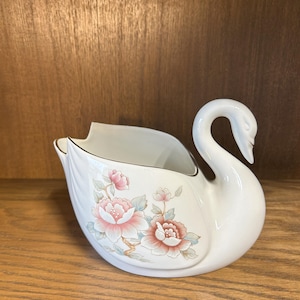 May include: A white ceramic swan planter with a floral design featuring pink and green flowers. The planter is shaped like a swan with its neck curved upwards.