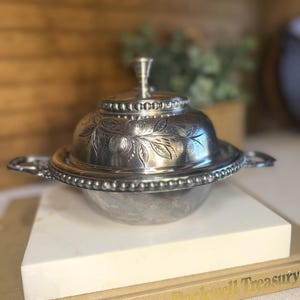 May include: A silver-toned metal butter dish with a domed lid featuring an engraved fruit and leaf pattern. The lid has a central finial knob, and the base includes a beaded rim edge with two decorative side handles for easy lifting and handling.
