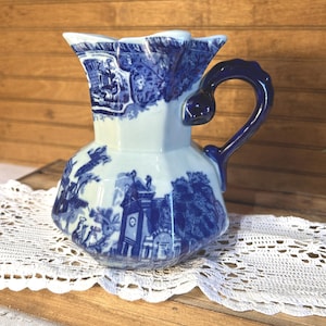 May include: A ceramic pitcher featuring a blue and white transferware pattern depicting architectural scenes and landscapes. The vessel has a faceted body, a wide pouring spout, and a solid dark blue curved handle attached to the side.