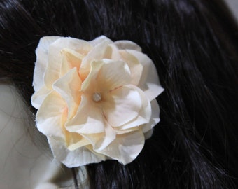 May include: A pale peach fabric flower hair clip. The flower is made of several layers of fabric and has a pearl-like center.