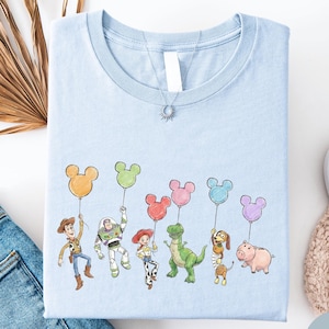 a light blue t-shirt with a playful design of cartoon characters holding up Mickey Mouse balloons, along with a pair of jeans and some other casual clothing items.
