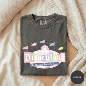 A gray t-shirt with a Disneyland logo and text, laid out on a bed with a white blanket.