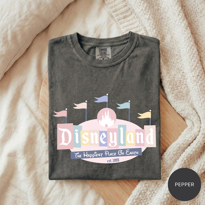 A gray t-shirt with a Disneyland logo and text, laid out on a bed with a white blanket.