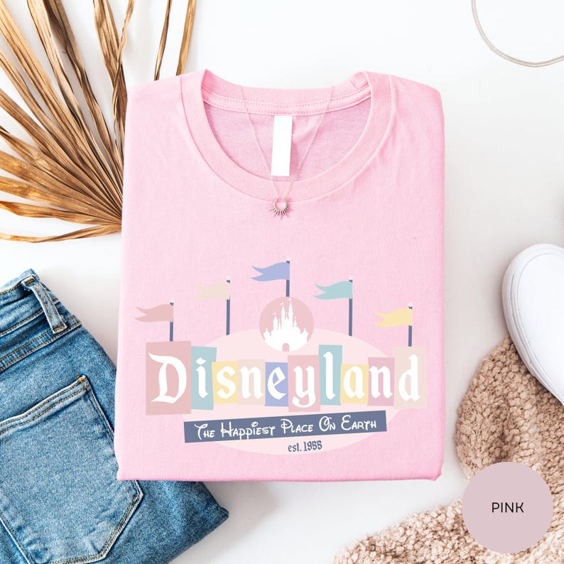 a pink t-shirt with a Disneyland logo, a pair of blue jeans, and some decorative elements such as a palm leaf and a beige sweater.