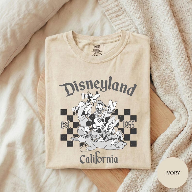 A beige t-shirt with a graphic design featuring Disney characters and text, laid out on a wooden surface.