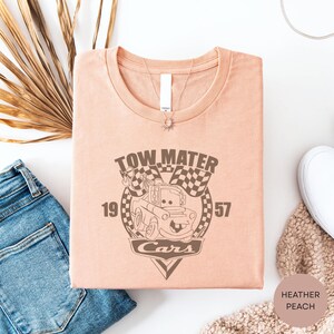a pink t-shirt with a graphic design of a tow truck and cars, along with the text "TOW MATTER" and "1957". The t-shirt is laid out on a white surface, accompanied by a pair of blue jeans, a white scarf, and a beige sweater.