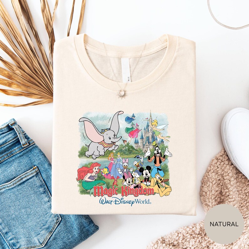 a white t-shirt with a colorful graphic of Disney characters and the text "Magic Kingdom" printed on it, along with a necklace and some jeans.