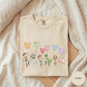 A beige t-shirt with a graphic design featuring characters from the Toy Story franchise, including Woody, Buzz Lightyear, and other beloved characters, along with various Disney-themed elements such as Mickey Mouse and Minnie Mouse balloons.