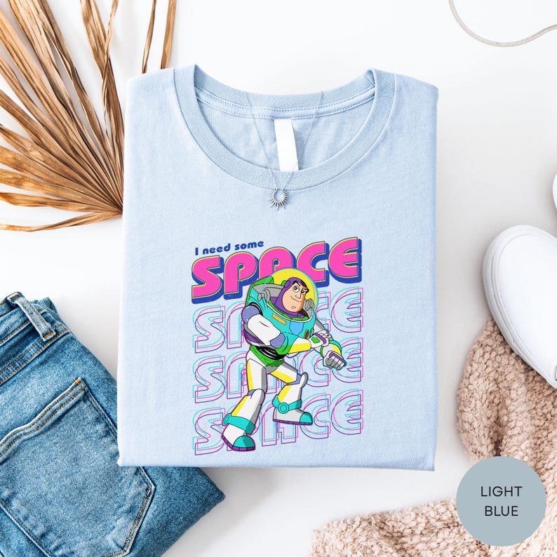 a light blue t-shirt with a graphic of a character from the animated film "Toy Story" and the text "I need some space" printed on it. The t-shirt is laid out on a white surface, along with a pair of jeans and some other clothing items.