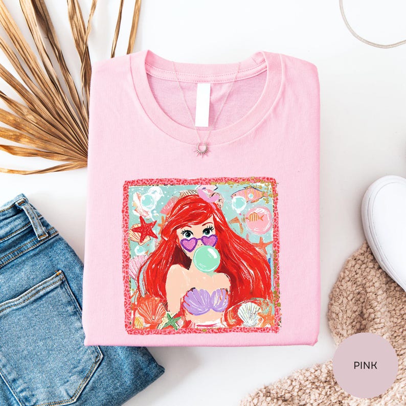a pink t-shirt with a colorful graphic of a mermaid, a pink flower, and a starfish. The t-shirt is folded and placed on a white surface, along with a pair of blue jeans and a beige sweater.