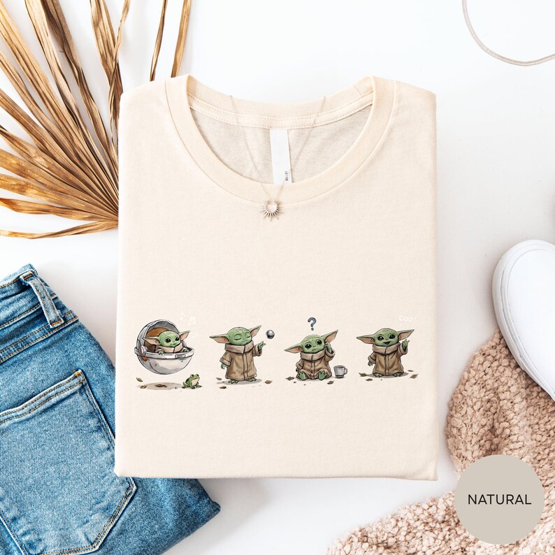 a t-shirt with a graphic design depicting four baby alien characters, each with a unique pose and expression. The t-shirt is laid out on a white surface, with a pair of blue jeans and a beige sweater visible in the background.