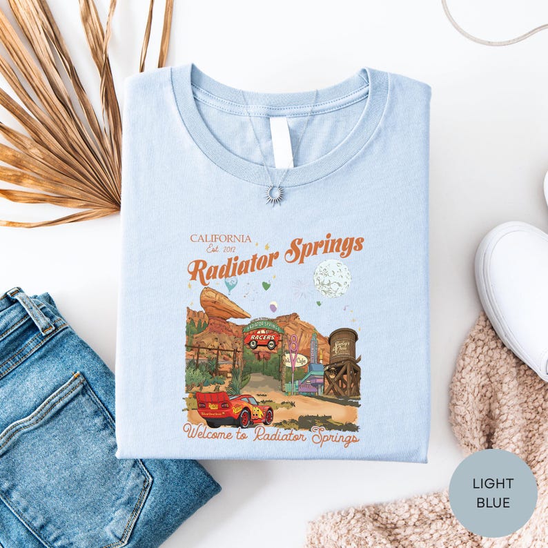 a light blue t-shirt with a graphic design depicting a car scene and the text "California Est 2012 Radiator Springs Welcome to Radiator Springs".