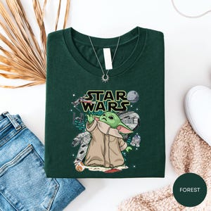 a green t-shirt with a Star Wars graphic, a pair of jeans, a necklace, and some beige and white clothing items, all laid out on a white surface.