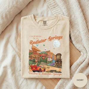 A t-shirt with a printed design depicting a scenic road leading to a red car, with the text "California Radiator Springs" and "Welcome to Radiator Springs" printed on it.