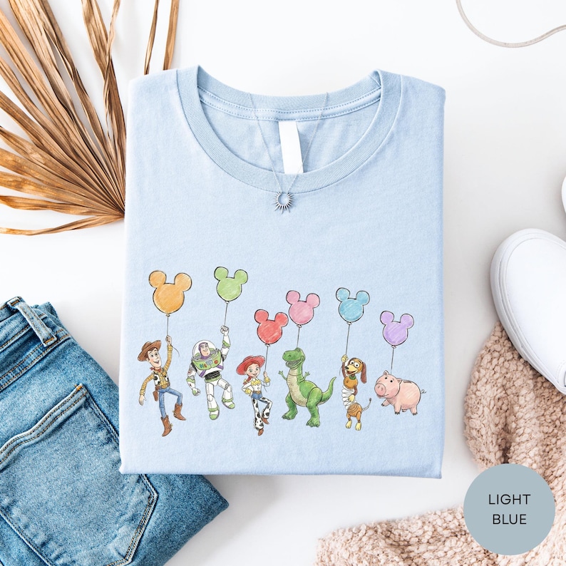 a light blue t-shirt with a playful design of cartoon characters holding up Mickey Mouse balloons, along with a pair of jeans and some other casual clothing items.