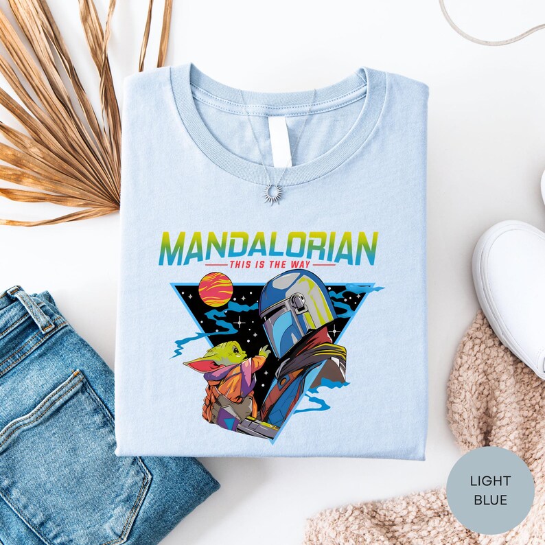 a light blue t-shirt with a colorful graphic design depicting a character from the Mandalorian TV series, along with the text "MANDALORIAN - THIS IS THE WAY". The t-shirt is laid out on a white surface, accompanied by a pair of jeans, a white sock, and a beige sweater.