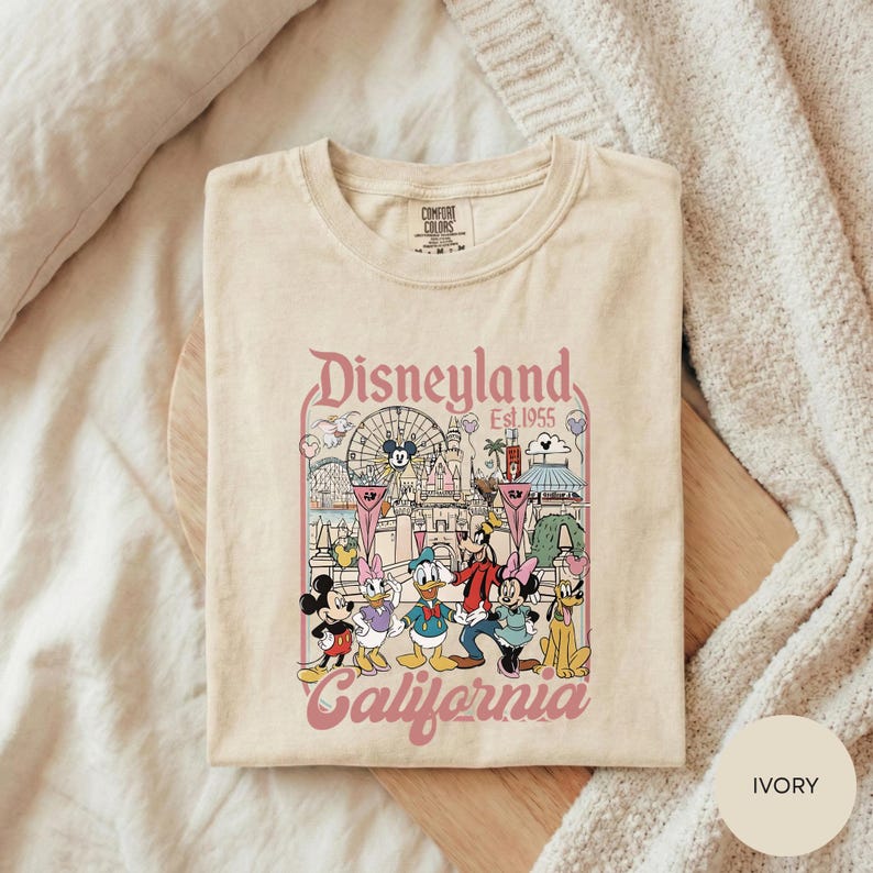 A beige t-shirt with a graphic design featuring Disney characters and the text "Disneyland Est. 1955" printed on it, laid out on a wooden surface.