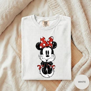 A white t-shirt with a Minnie Mouse graphic printed on it, placed on a wooden surface.
