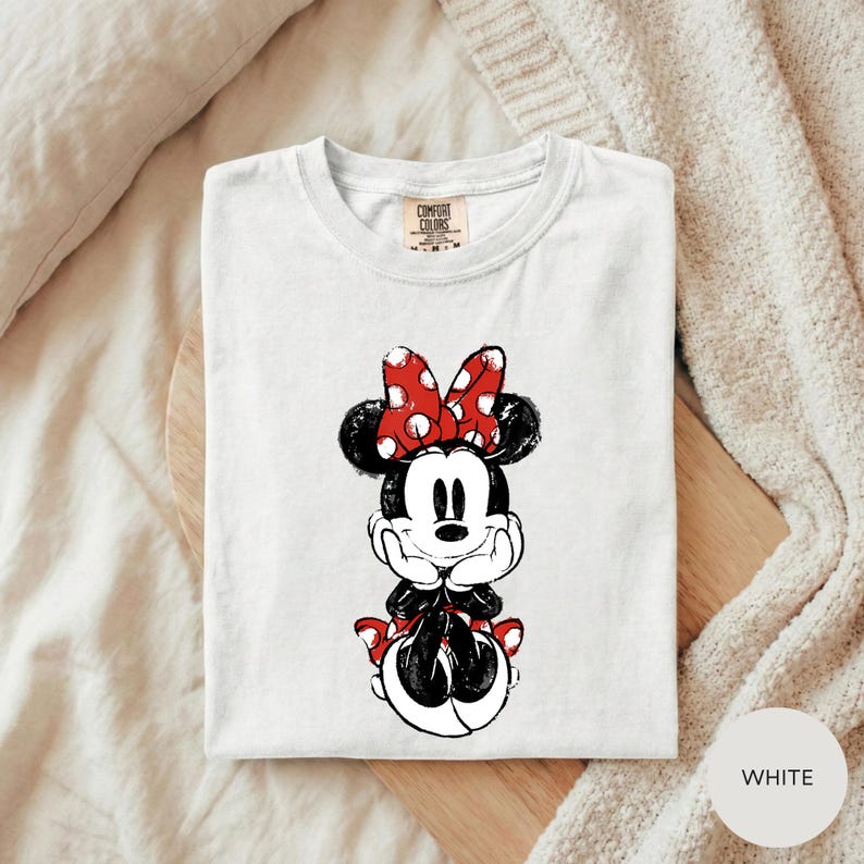 A white t-shirt with a Minnie Mouse graphic printed on it, placed on a wooden surface.