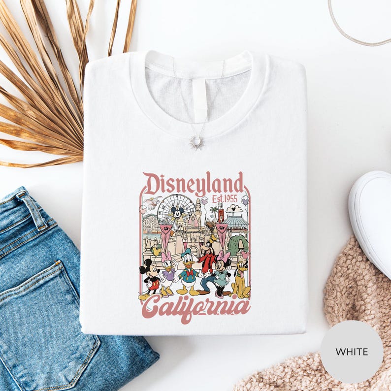 a white t-shirt with a Disneyland-themed graphic, including Mickey Mouse, Minnie Mouse, Donald Duck, and other iconic Disney characters, along with the text "Disneyland Est. 1955 California". The t-shirt is laid out on a white surface, with a pair of blue jeans and a beige sweater visible in the background.
