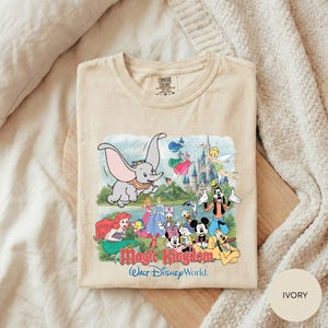 A t-shirt featuring a colorful illustration of Disney characters from the Magic Kingdom, including Dumbo, Minnie Mouse, Mickey Mouse, Donald Duck, and other iconic figures, set against a castle backdrop.