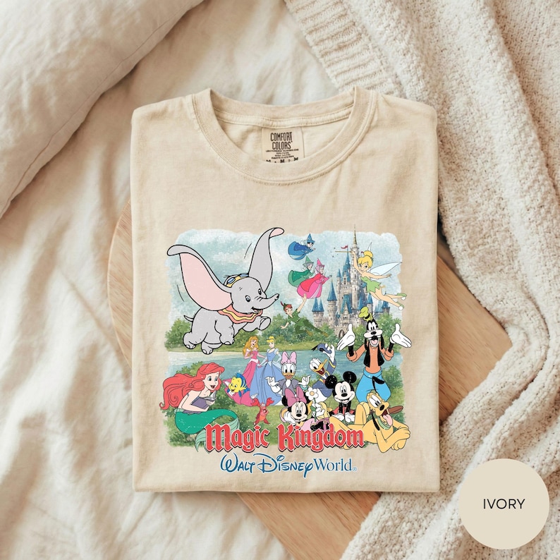 A t-shirt featuring a colorful illustration of Disney characters from the Magic Kingdom, including Dumbo, Minnie Mouse, Mickey Mouse, Donald Duck, and other iconic figures, set against a castle backdrop.