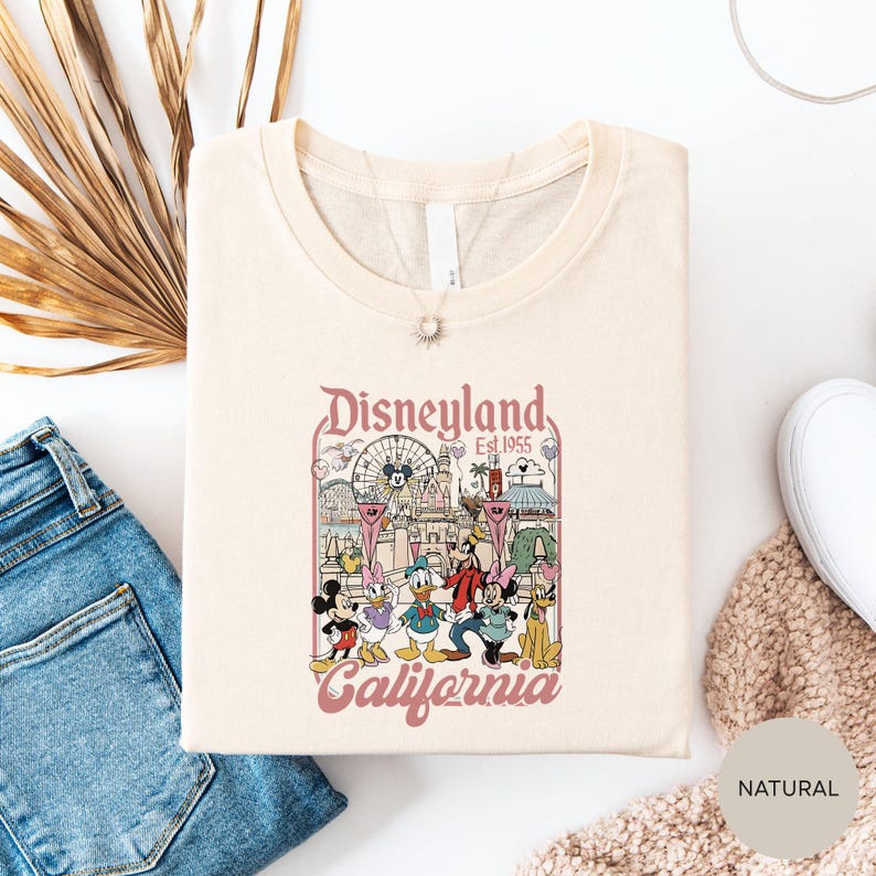 a white t-shirt with a Disneyland-themed graphic, including Mickey Mouse and other iconic characters, along with the text "Disneyland Est. 1955 California". The t-shirt is laid out on a white surface, with a pair of blue jeans and a beige sweater visible in the background.