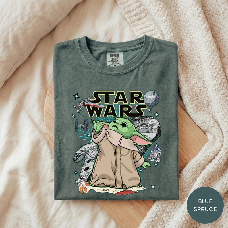 A green t-shirt with a graphic design featuring a young alien character, likely representing the character Baby Yoda from the Star Wars franchise, along with various Star Wars-themed elements such as a spaceship, a Death Star, and a Star Wars sign.