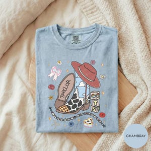 A blue t-shirt with a cartoon design featuring a cowboy hat, a bow, and various other icons, along with the text "TAYLOR" and "IT'S" printed on it.