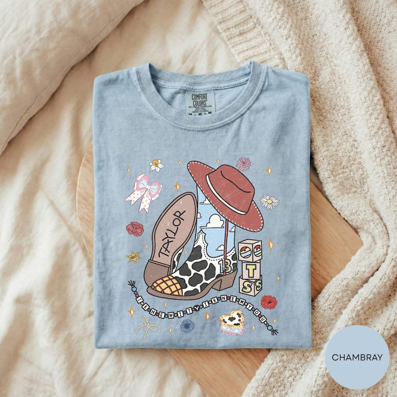 A blue t-shirt with a cartoon design featuring a cowboy hat, a bow, and various other icons, along with the text "TAYLOR" and "IT'S" printed on it.
