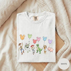 A white t-shirt with a graphic design featuring characters from the Toy Story movie, including Woody, Buzz Lightyear, and other beloved characters.
