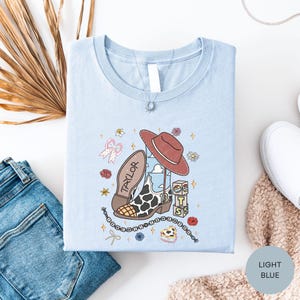 a light blue t-shirt with a cartoon character and various decorative elements, such as a cowboy hat, flowers, and stars, laid out on a white surface alongside a pair of jeans and a sweater.
