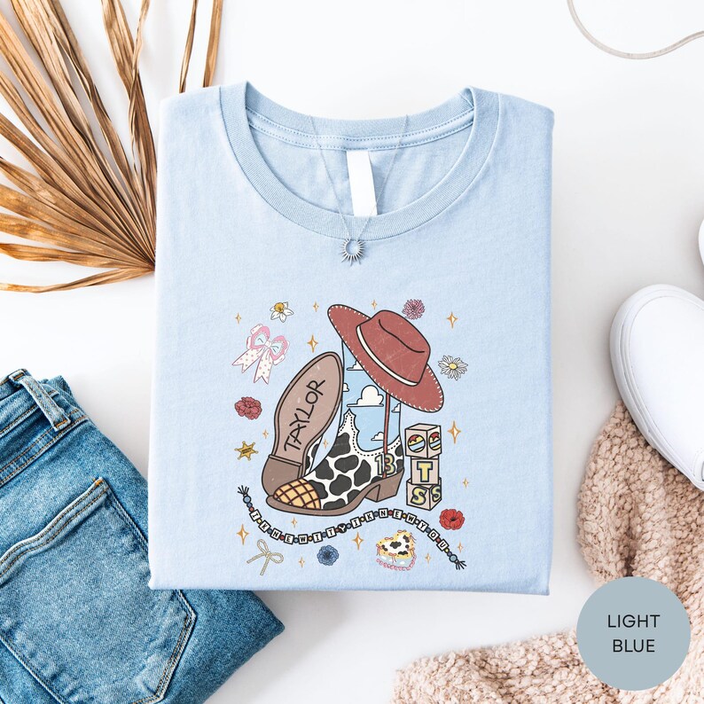 a light blue t-shirt with a cartoon character and various decorative elements, such as a cowboy hat, flowers, and stars, laid out on a white surface alongside a pair of jeans and a sweater.