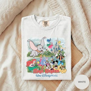 A white t-shirt with a colorful graphic design depicting various Disney characters from the Magic Kingdom, including Dumbo, Minnie Mouse, Mickey Mouse, Donald Duck, and other iconic figures.